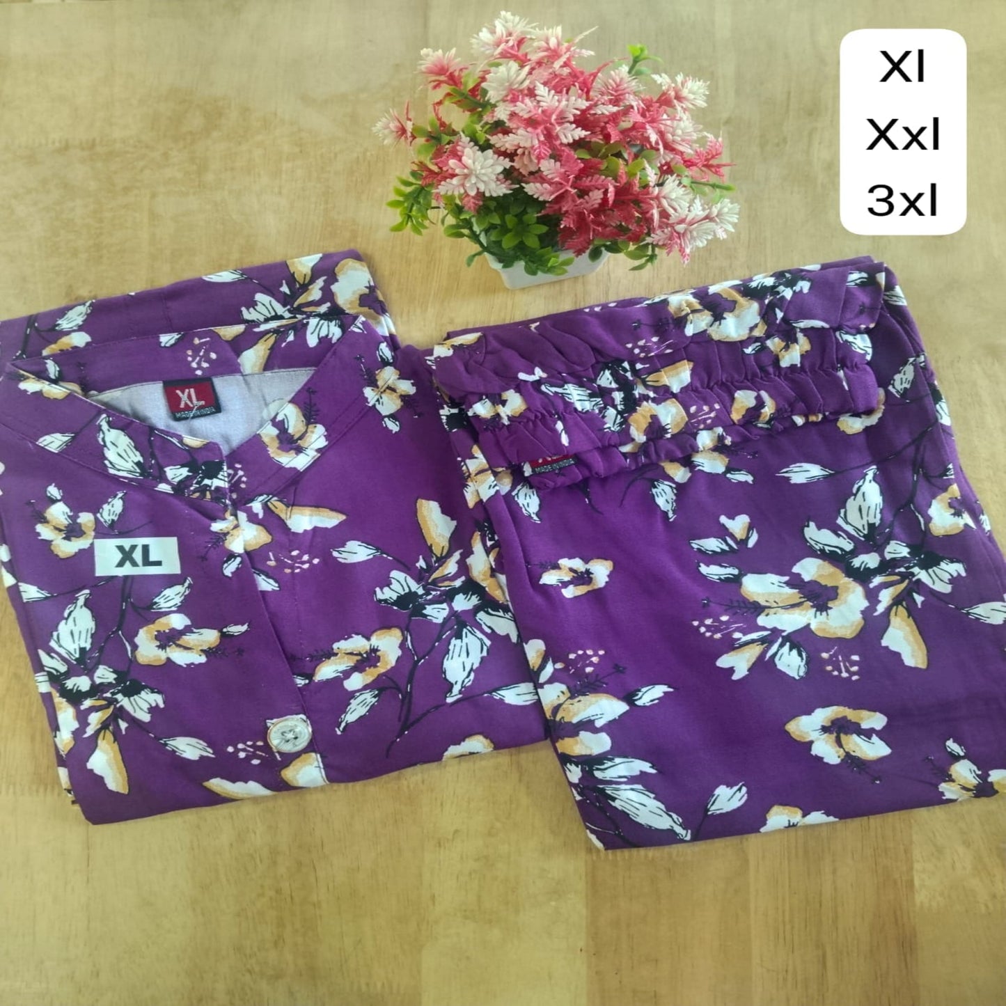 Purple floral pajama set with size labels on a wooden surface, accompanied by a small potted plant.